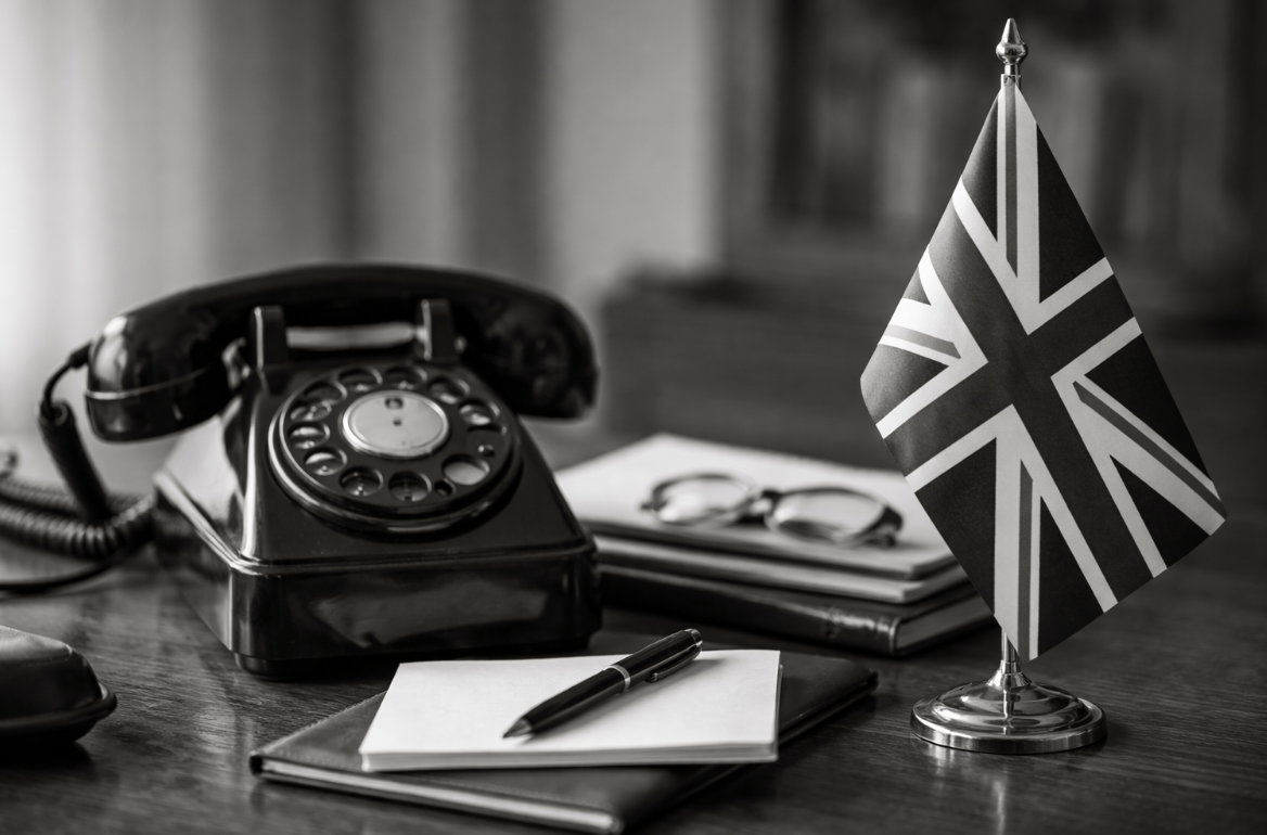 Bannière horizontale en noir et blanc montrant un téléphone ancien à cadran rotatif posé sur un bureau, à côté d’un petit drapeau britannique, avec un carnet, un stylo et des lunettes en arrière-plan légèrement flouté.